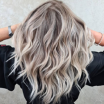 Клієнтка з хвилястим, попелясто-блонд волоссям AirTouch і темним корінням демонструє фарбування в салоні краси.