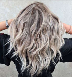 Клієнтка з хвилястим, попелясто-блонд волоссям AirTouch і темним корінням демонструє фарбування в салоні краси.